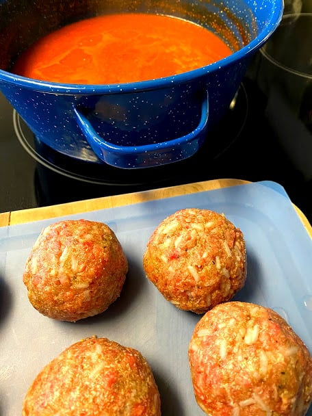 uncooked meatballs on plastic cutting board with pot of red salsa in the back drop