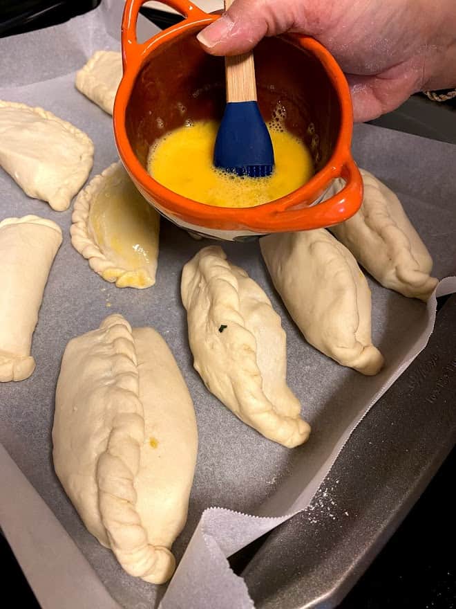 Unbaked Filled empanadas on lined baking sheet holding a small bowl of egg wash with pastry brush.